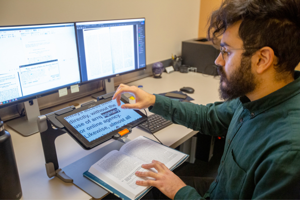 Man seated at a desk with two computer monitors operates a tablet displaying text with his right hand, while holding a book in place with his left hand