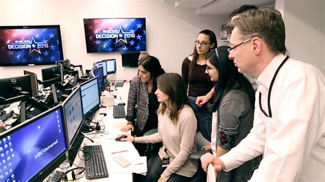 A man and a group of women gather around and study a monitor in a computer lab. Mounted against the wall beside them are two monitors displaying a "Decision 2016" logo graphic